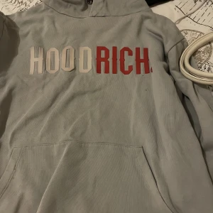 Ljusgrå HOODRICH hoodie med tryck - Säljer en ljusgrå hoodie från HOODRICH med stor vit och röd logga framtill. Klassisk känguruficka och huva. Perfekt för dig som gillar streetwear och vill sticka ut med en snygg och bekväm tröja. Köpt för 899, pris kan diskuteras 