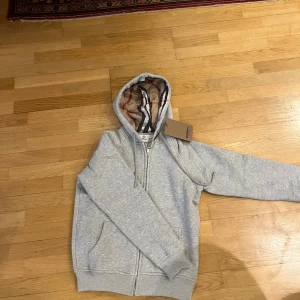 Grå hoodie från Burberry med dragkedja - Snygg grå hoodie från Burberry med klassiskt rutigt foder i huvan. Tröjan har dragkedja framtill, ribbade muddar och fickor på sidorna. Perfekt för dig som vill ha en stilren och exklusiv look med en touch av Burberrys ikoniska mönster. SÄLJES PGA FEL STORLEK ALDRIG ANVÄND