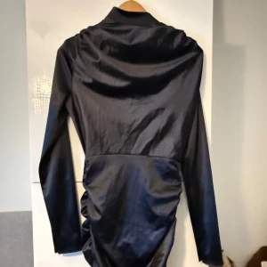 Svart långärmad satinklänning - Snygg svart klänning i satin med lång ärm och hög krage. Klänningen har en figurnära passform, rynkade detaljer längs sidorna och knäppning i nacken. Perfekt för dig som gillar en elegant och stilren look. Använd 1 gång. 