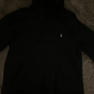 Svart hoodie från Polo Ralph Lauren - Svart hoodie från Polo Ralph Lauren med klassisk vit broderad logga på bröstet. Tröjan har huva med snörning och en stor magficka framtill. Perfekt för dig som gillar stilren och enkel streetwear.