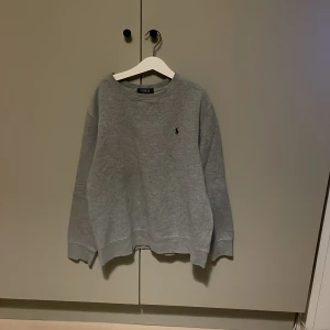 Grå sweatshirt från Polo Ralph Lauren - Klassisk grå sweatshirt från Polo Ralph Lauren med rund halsringning och broderad marinblå logga på bröstet. Tröjan har ribbade muddar vid ärmslut och nederkant, perfekt för en chill och stilren look | priset kan alltid diskuteras!