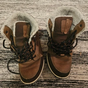 Bruna vinterkängor från Ecco - Snygga bruna kängor från Ecco med grov sula och snörning. Skorna är fodrade med mjukt, ljust foder för extra värme. Tillverkade i skinn och mocka med svarta och orange detaljer på sulan. Perfekta för kalla dagar och outdoor vibes.