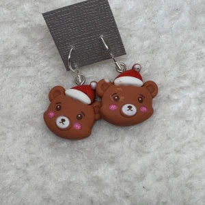 Söta björnörhängen med tomteluva - Supergulliga örhängen med bruna björnhuvuden som har röda och vita tomteluvor. Björnarna har rosa kinder och små detaljerade ansikten, perfekta för att ge julkänsla till din look. Hängande modell med metallkrokar.