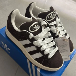 Adidas Campus mörkbruna - Adidas Campus 00s sneakers i mörkbrunt mocka med vita detaljer och klassiska tre ränder på sidan. Skorna har tjocka vita snören, rund tå och vit sula. Helt ny aldrig använt den prislapp på samt kartongen till skorna 