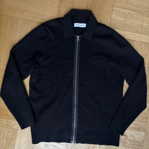 Svart kofta med dragkedja Samsøe Samsøe - Svart kofta från Samsøe Samsøe med hel dragkedja framtill och klassisk krage. Tröjan har en enkel bröstficka och är långärmad. Perfekt för lager-på-lager och stilren look.