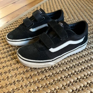 Svarta Vans sneakers med kardborre - Svarta Vans sneakers med klassisk vit sidorand och vit sula. Skorna har två breda kardborreband för enkel påtagning och en låg profil. Skorna är slitna, men håller ett tag till.