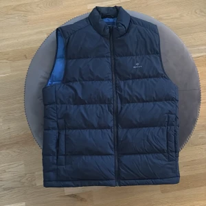 Mörkblå dunväst från GANT - Snygg mörkblå dunväst från GANT med quiltad design och hög krage. Västen har dragkedja framtill och en blå insida som ger en fräsch kontrast. Perfekt för lager-på-lager och stilren look.