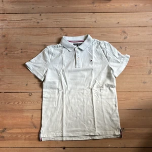 ljusblå pikétröja från Tommy Hilfiger 164 cm - Klassisk vit pikétröja från Tommy Hilfiger med korta ärmar och liten broderad logga på bröstet. Tröjan har krage, två knappar och diskreta ränder på insidan av kragen. Perfekt till jeans eller shorts för en clean look. Nypris 500 kr.
