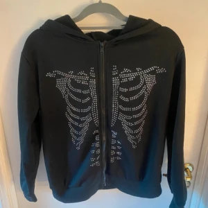 Svart zip hoodie med skelettmönster - Svart hoodie med dragkedja och huva. Framsidan har ett coolt skelettmönster i silvriga nitar som ger en edgy vibe. Perfekt för dig som gillar unika detaljer och streetstyle. Långärmad och skön att bära.