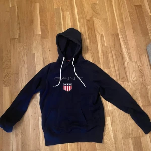 Mörkblå hoodie från GANT - Säljer en marinblå hoodie från GANT med logga framtill. Tröjan har huva med vita snören och en stor magficka framtill. Nypris:1000kr