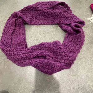 Lila stickad halsduk - Mysig och chunky lila halsduk i grovstickat garn. Halsduken är rektangulär och extra bred, perfekt att vira flera varv runt halsen för maximal värme. Färgen är en djup lila som verkligen poppar och passar perfekt till kalla dagar.