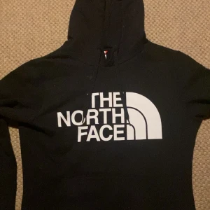Svart hoodie från The North Face - Svart hoodie från The North Face med stor vit logga framtill och mindre logga på ryggen. Klassisk huva med snörning och känguruficka. Mjuk bomullsblandning och ribbade muddar vid ärmslut och nederkant. Perfekt för chill dagar eller när du vill ha en clean streetstyle.