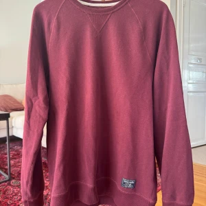 Vinröd sweatshirt från Abercrombie - Vinröd sweatshirt från Abercrombie & Fitch med rund halsringning och långa ärmar. Tröjan har en klassisk passform och en liten tygpatch nertill. Perfekt för dig som gillar enkel och stilren streetwear.