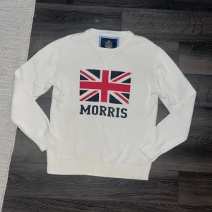 Tröja Morris - Snygg vit stickad tröja från Morris med Union Jack-flagga och Morris-logga på bröstet. Tröjan har rund halsringning, ribbade muddar och är långärmad. Perfekt för dig som gillar klassisk brittisk stil och vill sticka ut. Storlek XL.  Men jag tycker den sitter som L! Men utgå från måtten för att vara säker :)Axelbredd 50cm, Längd 71cm. JAG SKICKAR INGA FLER BILDER PÅ NÄR JAG BÄR PLAGGET:) 