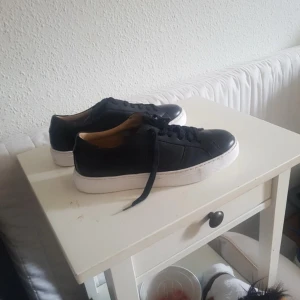 Svarta sneakers från Marstrand strl 40 - Snygga svarta sneakers från Marstrand med vit platt sula och klassisk snörning. Skorna har en stilren och enkel design med rund tå och är tillverkade i skinn för en schysst look. Perfekta för dig som gillar minimalistisk stil och vill ha något som funkar till det mesta. 