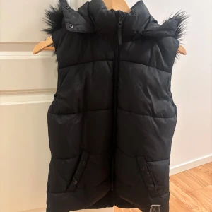 Svart dunväst med fuskpäls från H&M - Svart dunväst från H&M med huva och fuskpälsdetalj runt kanten. Västen har dragkedja framtill, två fickor med knappar och quiltad design. Perfekt för lager-på-lager och snygg till höst- och vinteroutfits.