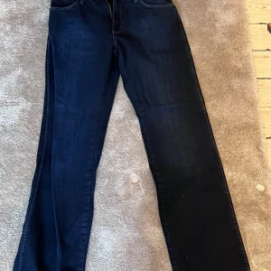Mörkblå Wrangler jeans W31 L34 - Snygga mörkblå jeans från Wrangler i klassisk modell med raka ben och ikoniska bakfickor med W-detalj. Tillverkade i 100% bomull för en skön känsla. Perfekta för dig som gillar tidlös denimstil. ALDRIG ANVÄNDA