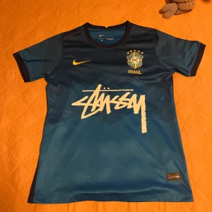 Blå Brasil Nike x Stüssy fotbollströja - Unik blå Brasilien fotbollströja från Nike i samarbete med Stüssy. Tröjan har korta ärmar, gul Nike-logga och Brasil-märke på bröstet samt Stüssy-tryck framtill. Tillverkad i Dri-FIT-material för bästa komfort. Perfekt för dig som gillar streetwear och fotboll.