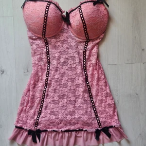 Rosa spetsklänning med volanger och rosetter - Supersöt rosa kort klänning i spets med vadderade kupor, svarta rosetter och dekorativa band framtill. Volangkant nertill och flera volanger på ryggen. Justerbara axelband och öppen rygg för extra detalj. Perfekt för dig som gillar romantisk och lekfull stil.