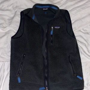 Mörkgrå fleeceväst från Patagonia - Mysig mörkgrå fleeceväst från Patagonia med blå detaljer vid dragkedjorna och insidan av kragen. Västen har tre fickor med dragkedja och en hög krage. Perfekt för lager-på-lager och outdoor-aktiviteter. Snygg, enkel design med diskret logga på bröstet.