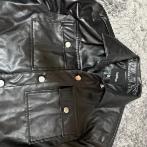 Svart skinnjacka från Boohoo - Snygg svart skinnjacka från Boohoo med croppad passform. Jackan har stora bröstfickor med lock och silvriga knappar framtill. Tillverkad i syntetmaterial med en glansig finish och klassisk krage. Perfekt för dig som vill ha en edgy look.