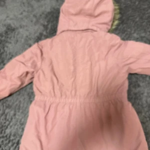 Rosa parkajacka med fuskpäls och dragkedja - Parkajacka i ljusrosa med mysig vit fuskpäls på insidan och huva med pälsdetalj. Jackan har dragkedja framtill, knappar vid halsen och praktiska fickor med knappstängning. Justerbar midja med snörning för snygg passform.