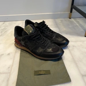 Valentino Rockrunner svarta sneakers - Svarta Valentino Rockrunner sneakers med stjärnmönster i mocka och skinn, vinröd häl med nitar och stjärndetalj i silver. Snygga svarta snören och platt sula i gummi. Märkeslogga på plösen och yttersulan. Perfekt för dig som gillar exklusiv streetstyle.