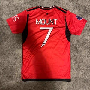 Manchester United Mount 7 Adidas tröja - Manchester Uniteds officiella hemmatröja från Adidas med Mount och nummer 7 på ryggen. Röd med svarta och vita detaljer, klubbmärke på bröstet och Premier League-logga på ärmen. Tillverkad i lätt och ventilerande polyester med snyggt mönster i tyget.