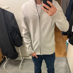 Ljusgrå kofta med dragkedja dobber  - Säljer en stilren ljusgrå kofta med hel dragkedja och ribbade muddar. Tröjan har hög krage och är perfekt för lager-på-lager. Enkel och clean design som passar till jeans eller chinos. Skick 9/10