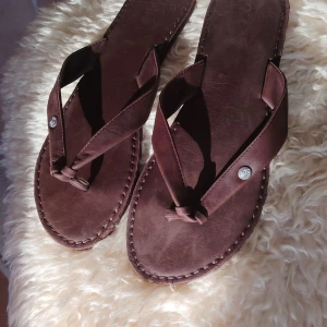 Abercrombie & Fitch sandaler i äkta skinn  - Snygga bruna sandaler från Abercrombie & Fitch i mjukt skinn. Klassisk flipflop-modell med breda remmar och dekorativa metallknappar. Sulan är platt och har synliga sömmar runt kanten för en stilren look. Perfekta för varma dagar.