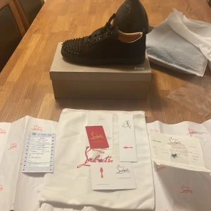 Christian Louboutin svarta sneakers med nitar - Säljer ett par svarta sneakers från Christian Louboutin med coola svarta nitar på tån och klassisk röd sula. Skorna har snörning och är tillverkade i skinn och textil. Kommer med originalkartong, dustbag och äkthetsbevis. Perfekta för dig som vill sticka ut.