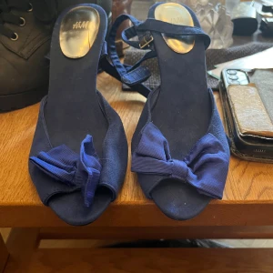 Blå pumps med rosett från H&M - Snygga blå pumps från H&M med smal klack och öppen tå. Skorna har en dekorativ rosett framtill och smala remmar med spänne runt ankeln. Materialet är mockaliknande och färgen är djupblå, vilket ger en elegant och trendig look.