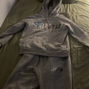 Grå tracksuit från Trapstar med tryck - Säljer en äkta tracksuit från Trapstar med stor logga och text på bröstet. Tröjan har huva med snörning och en stor magficka. Perfekt för dig som gillar streetwear och vill ha en skön och avslappnad stil. Dm för fler bilder 