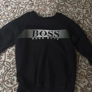 Svart sweatshirt från Hugo Boss - Svart sweatshirt från Hugo Boss med stort silvrigt logotryck och prickigt mönster över bröstet. Tröjan har rund halsringning och långa ärmar. Perfekt för en stilren och sportig look.