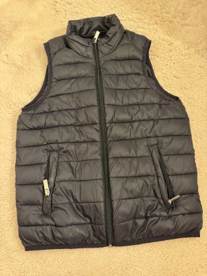 Svart quiltad dunväst med dragkedja - Svart dunväst med quiltad design och hög krage. Västen har två sidofickor med dragkedja och en hel dragkedja framtill. Perfekt för lager-på-lager och har en stilren look som funkar till många outfits.