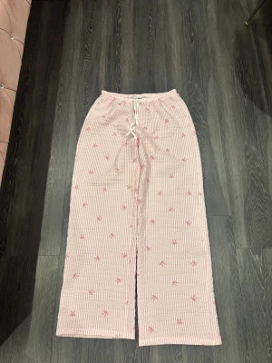 Rosa randiga byxor med rosetter - Säljer ett par ljusrosa byxor med vita ränder och små broderade rosetter i rosa. Byxorna har snörning i midjan och är vida i modellen. Perfekta för en chill och söt look.