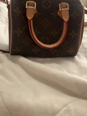 Louis Vuitton monogram handväska - Säljer en klassisk handväska från Louis Vuitton med ikoniskt monogrammönster i mörkbrunt och beige. Väskan har två handtag i ljust skinn med röda detaljer och dragkedja upptill. Perfekt storlek för det viktigaste och riktigt snygg design.