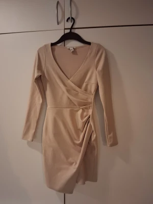 Beige omlottklänning från NLY One XS - Snygg beige kortklänning från NLY One i omlottmodell med v-ringning och långa ärmar. Klänningen har draperad detalj i midjan som ger en cool och trendig look. Perfekt för dig som gillar stilrena och enkla plagg med en twist.