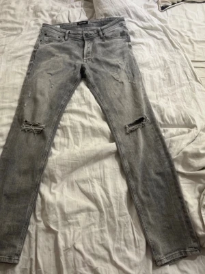 Grå slitna jeans Jack & Jones Glenn - Säljer ett par grå jeans från Jack & Jones, modell Glenn. Jeansen har skinny fit, slitningar och hål på båda knäna för en cool, avslappnad look. Klassisk femficksmodell med dragkedjegylf och snygga detaljer på bakfickorna.