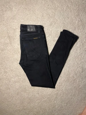 Svarta Nudie Jeans 30/30 - Svarta jeans från Nudie Jeans med klassisk femficksdesign och svarta sömmar. Jeansen har en smal passform och är tillverkade i stretchigt denim för extra komfort. Perfekt för dig som gillar en stilren och modern look.
