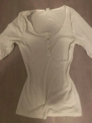 Vit ribbad topp med knappar från H&M - Säljer en vit ribbad topp från H&M med rundad halsringning och fyra dekorativa knappar framtill. Toppen har trekvartsärm och är figurnära i modellen, perfekt för dig som gillar en clean och enkel look.