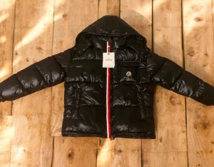Svart Moncler pufferjacka med huva - Svart pufferjacka från Moncler med glansig finish, huva och rödvit dragkedja framtill. Jackan har en ficka med Moncler-logga på bröstet och två sidofickor. Perfekt för kalla dagar och har en riktigt snygg streetstil.