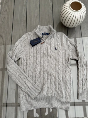 Grå stickad half zip-tröja Polo Ralph Lauren - Snygg grå kabelstickad tröja från Polo Ralph Lauren med half zip och ribbade muddar. Tröjan har lång ärm, hög krage och klassisk broderad logga på bröstet. Perfekt för dig som gillar stilren och tidlös design.