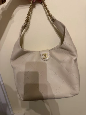 Beige quiltad axelväska  - Säljer en beige axelväska från  med klassiskt quiltat mönster och guldfärgade kedjedetaljer. Väskan har den ikoniska C-loggan i guldaktig på framsidan är också quiltad. 