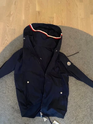  Moncler jacka - Snygg mörkblå vindjacka från Moncler med huva och dragsko, silverfärgade knappar och dragkedja framtill. Perfekt för dig som gillar exklusiv look. Nästan helt ny använd max 2 gånger, skick 9/10.  Det står storlek M i jackan men är S. Givetvis äkta, NFC kod funkar och allt!