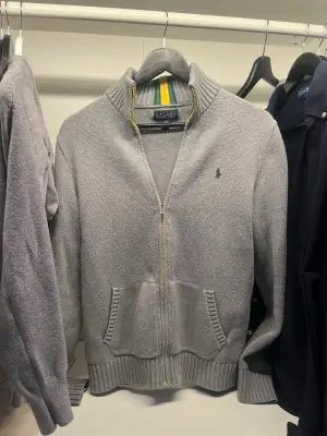 Grå stickad kofta med dragkedja från Polo Ralph Lauren. Har ribbad krage med gröna och gula ränder, två fickor fram och broderad grön logga på bröstet. Pris 699