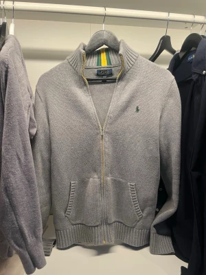 Grå stickad kofta från Polo Ralph Lauren - Grå stickad kofta med dragkedja från Polo Ralph Lauren. Har ribbad krage med gröna och gula ränder, två fickor fram och broderad grön logga på bröstet. Pris 699