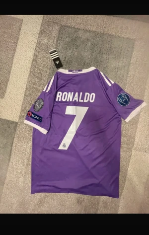 lila Real Madrid Ronaldo 7  - Bilderna visar en lila Real Madrid Ronaldo 7 tröja från Adidas, med Champions League och FIFA-märken, men inga skor finns med.