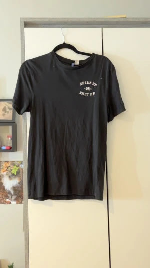 Svart t-shirt med tryck 'Speak Up Or Shut Up' - Svart t-shirt från Gildan med vit text 'SPEAK UP OR SHUT UP' tryckt på bröstet och i större format på ryggen. Klassisk passform med rund hals och korta ärmar. Perfekt statement-plagg för dig som vill sticka ut.