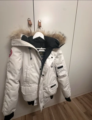 Vit dunjacka Canada Goose med päls - Säljer en snygg vit dunjacka från Canada Goose med fluffig äkta päls på huvan. Jackan har svarta muddar, dragkedja och knappar framtill samt flera fickor. Klassisk patch på ärmen och cool, oversized look. Perfekt för kalla vinterdagar.